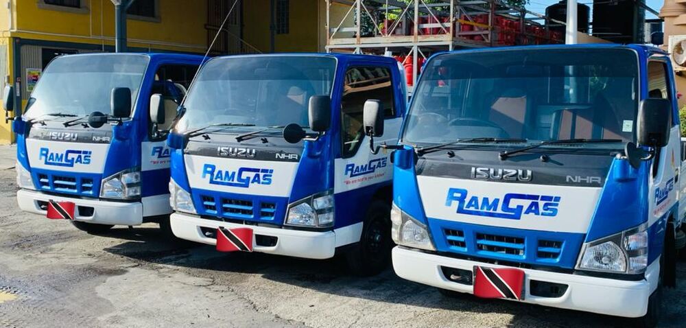 Three blue and white RamGas Isuzu cooking gas delivery trucks lined up, each with a Trinidad and Tobago flag on the bumper