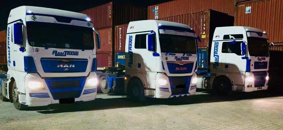 Three Ramtrans MAN TGX tractor units with headlights on at night, parked in front of stacked shipping containers